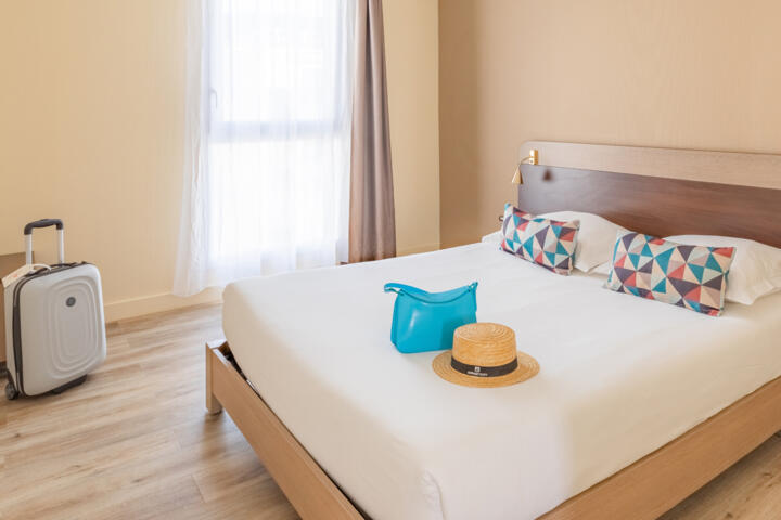 Habitación de hotel de la gama clásica con una cama doble grande, cojines coloridos, una maleta blanca con ruedas y un bolso turquesa con un sombrero de paja encima, evocando una atmósfera acogedora para los viajeros.
